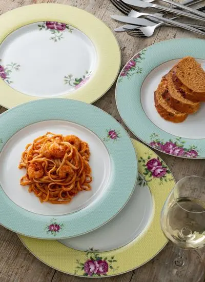 A dinner table setup featuring a plate of spaghetti with tomato sauce, slices of bread on a side plate, empty floral plates, utensils, and a glass of white wine.